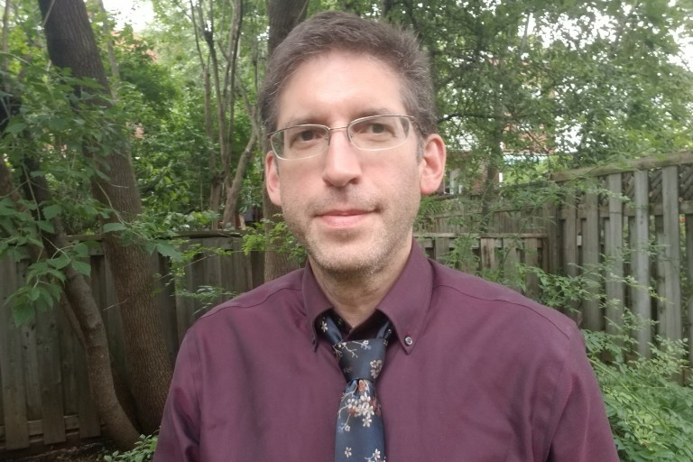 A man with a tie and glasses stands in front of a fence, with some greenery in the background.
