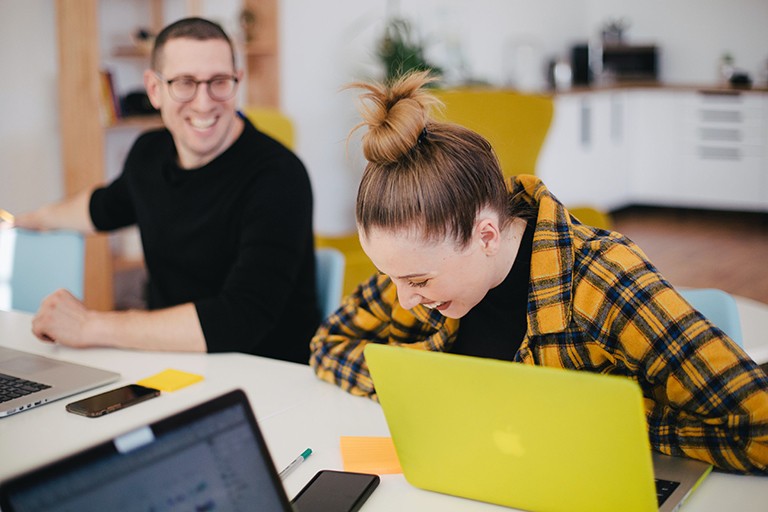 A laughing woman and man sitting at desk in front of laptops.