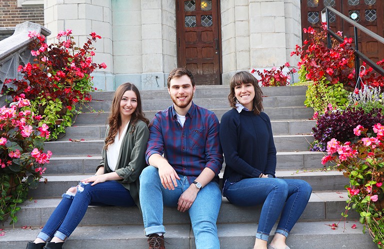 Three young people — two women and one man — sitting on steps in front of a building.