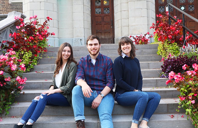 Three young people - two women and one man - sitting on steps in front of a building.