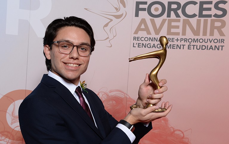 Young, smiling man with glasses, wearing a suit and holding up an award (a figure, arms outstretched, in gold).