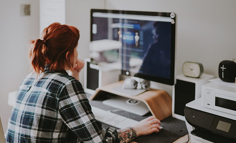 Young woman with red hair, an arm tattoo and a checkered shirt sitting at a computer.