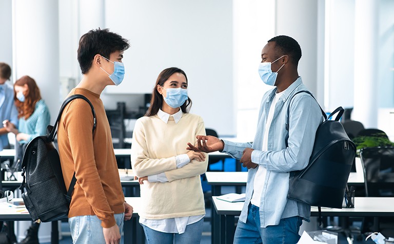 Young people wearing masks, standing around and talking in a classroom setting.