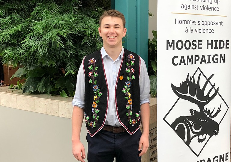 Young, smiling man wearing a colourful vest and standing beside a poster that says, "Moose Hide Campaign."