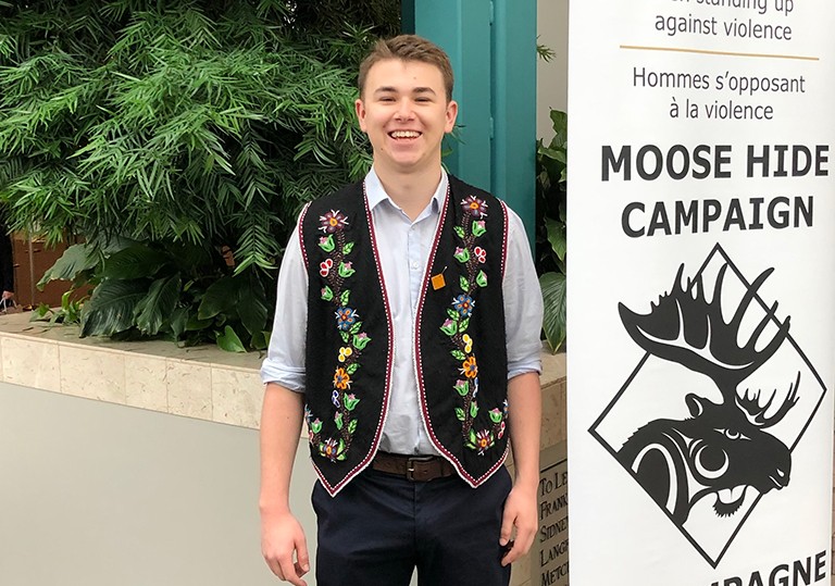 Aiden Cyr: “As an Indigenous person, you are immersed in politics from the get-go.” Young, smiling man wearing a colourful vest and standing beside a poster that says, "Moose Hide Campaign."