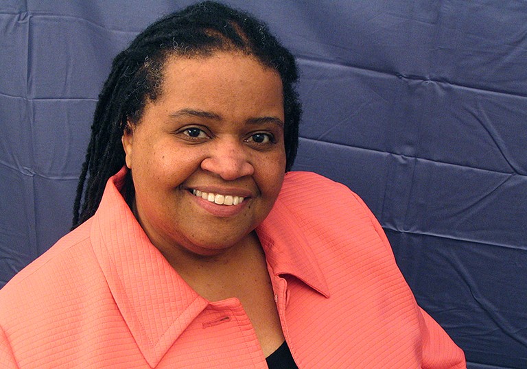 Picture of a smiling Black woman with a coral coloured jacket.