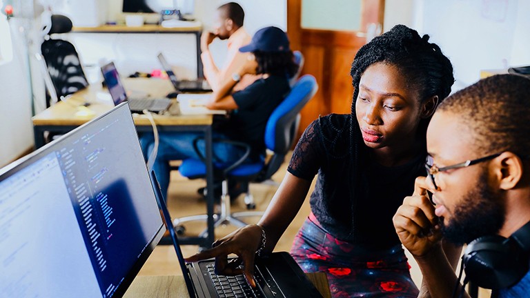 Pictured: A Black woman and Black man sitting in front of a desk-top computer, and the woman is pointing to something on the screen.