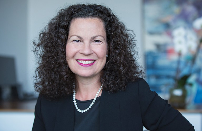 Smiling woman with long, dark, curly hair, wearing a black top and jacket.