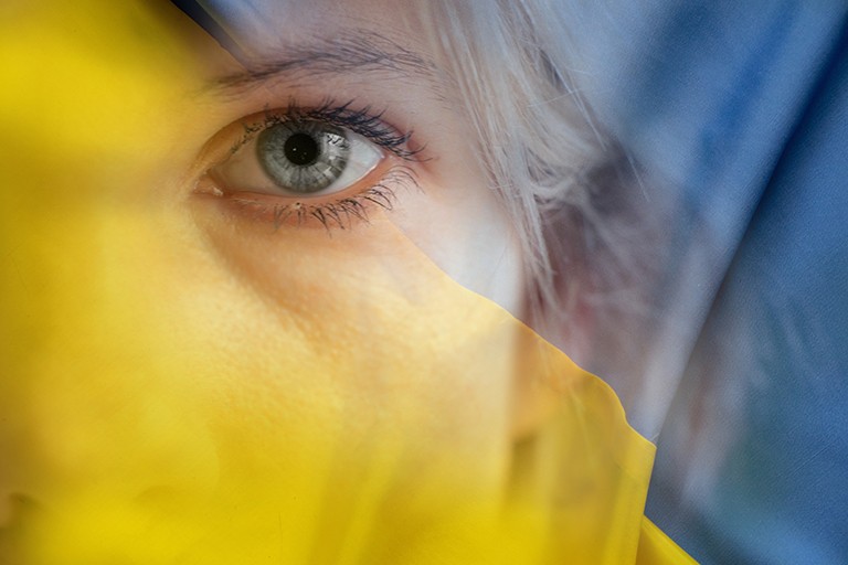 Olena Zamkovaya is a Concordia student of Ukrainian origin currently doing on-the-ground refugee support work at the Ukraine-Hungary border. Close-up on the blue eye of a young woman, with a see-through yellow scarf over half her face.