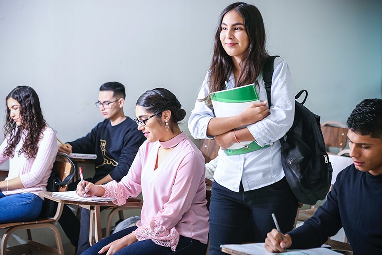 A group of university students sitting at desks and writing, with one girl standing and holding a large textbook in her arms.