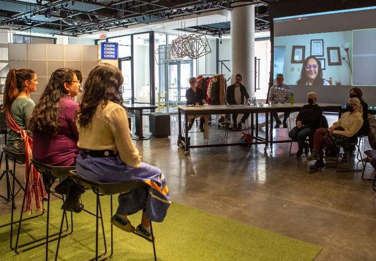 Miywâcimo! storytelling competition Three women on stools facing judges table, audience members and screen projection of Zoom participants