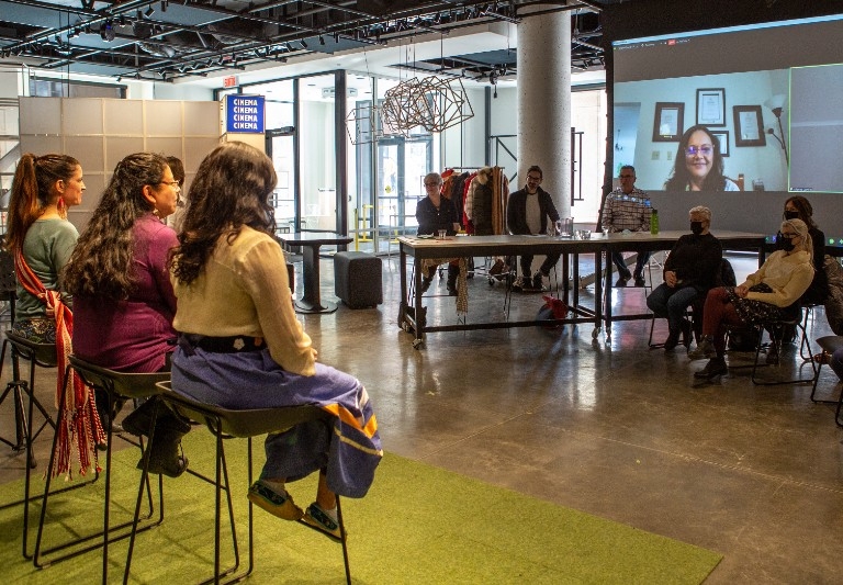 Three women on stools facing judges table, audience members and screen projection of Zoom participants