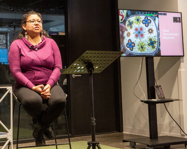 Amanda Shawayahamish K.Wynn photo by Lisa Graves Woman wearing purple sweater, sitting in stool, with screen presentation slide depicting beadwork