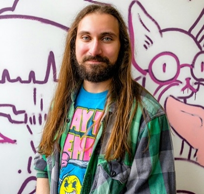 Young smiling man with long, straight blond hair and a colourful shirt.