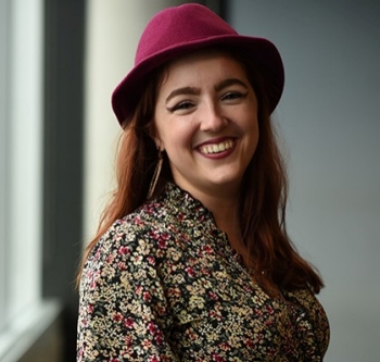 Young smiling woman, with long dark red hair, a red hat and a patterned shirt.