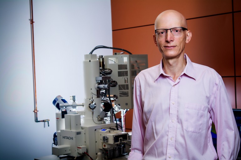 Man with glasses and a pink shirt standing in front of lab equipment.