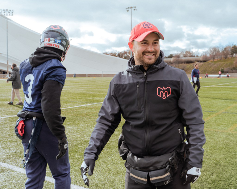 Two men standing on a football field, one is a player and the other is a trainer.