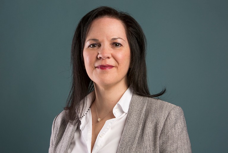 Smiling woman with long, dark hair, wearing a white shirt with a beige dress jacket