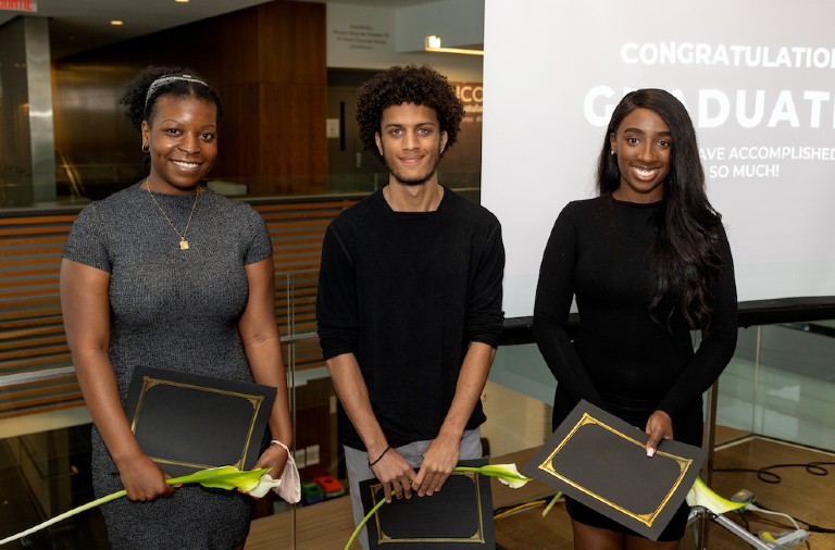 Three students holding their diplomas and a white flower