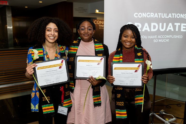 Three female students smiling holding their diplomas and a white flower