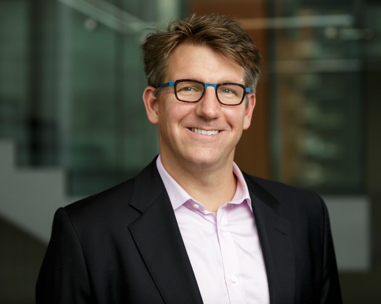 Headshot of smiling man wearing black-and-blue glasses, black blazer and light-purple collared shirt