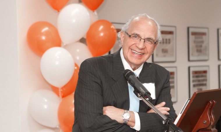 Man in suit smiles at microphone with arms crossed, white and orange balloons in background