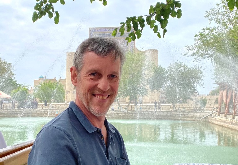 Richard Foltz: A smiling man sitting in front of a fountain on a sunny day.