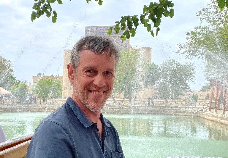 A smiling man sitting in front of a fountain on a sunny day.