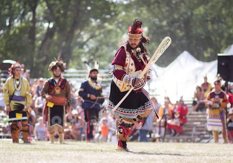 A group of Indigenous men dressed in traditional clothes dance in an outdoor, open area.