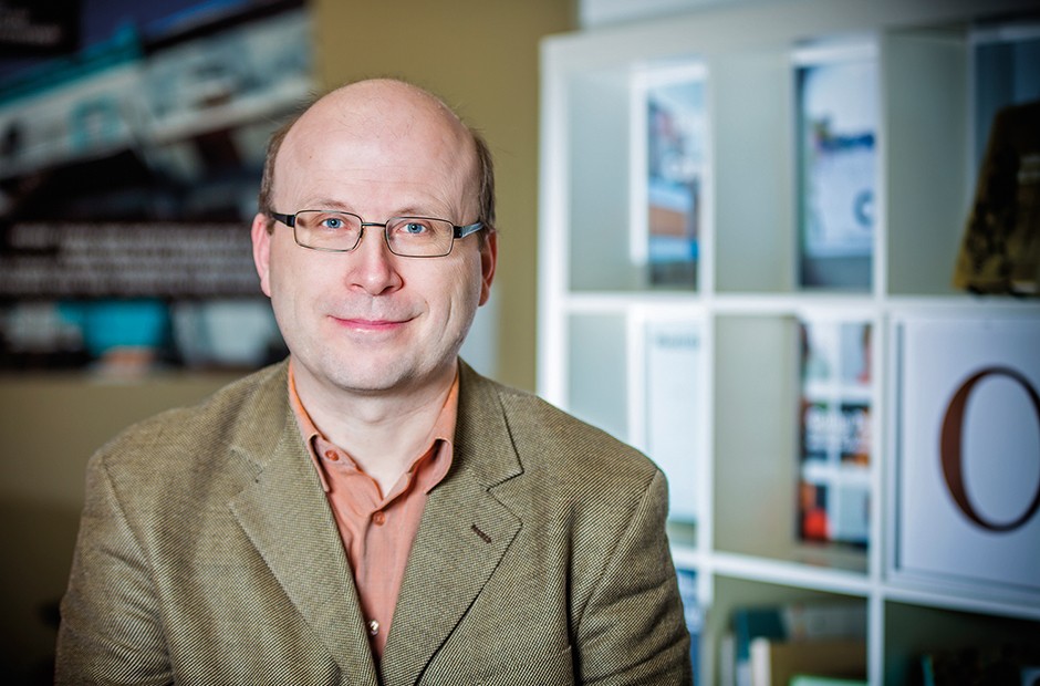 Bald, smiling man wearing spectacles and a brown suit jacket with an apricot-coloured shirt.