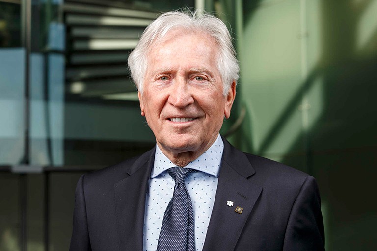 Older man with silver-white hair standing in front of a glass wall, wearing a suit and tie.