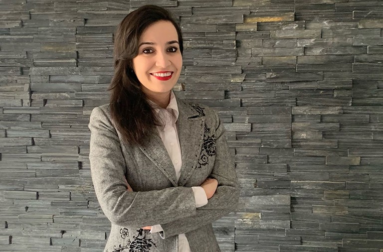 Young, smiling woman with long, dark hair, red lipstick and wearing a grey blazer with a white shirt.