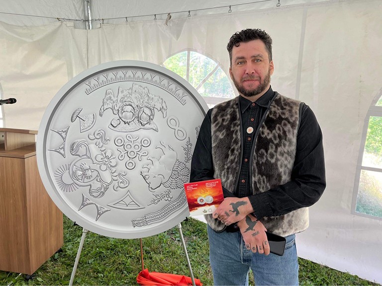 Jason Sikoak stands next to the Truth and Reconciliation keepsake medallion that he helped design. A bearded man standing next to the sculptural representation of a silver coin