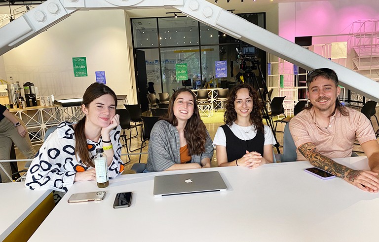 A group of four smiling young people (three women and one man) sitting at a table in a gallery space and looking at the camera