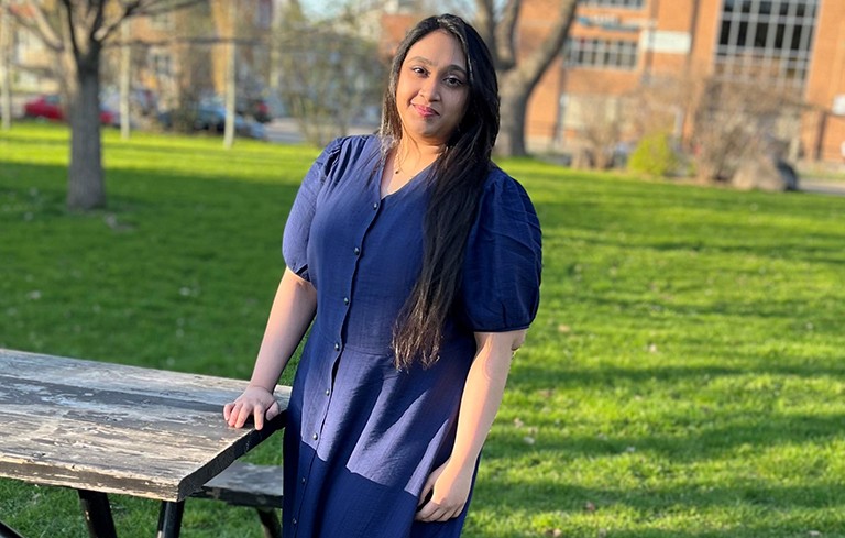 A young woman with long, dark hair, wearing a blue dress and standing outside with her hand on a picnic table