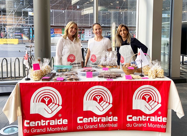 Three women standing behind a table with sweet treats and a banner saying, "Centraide du Grand Montreal"