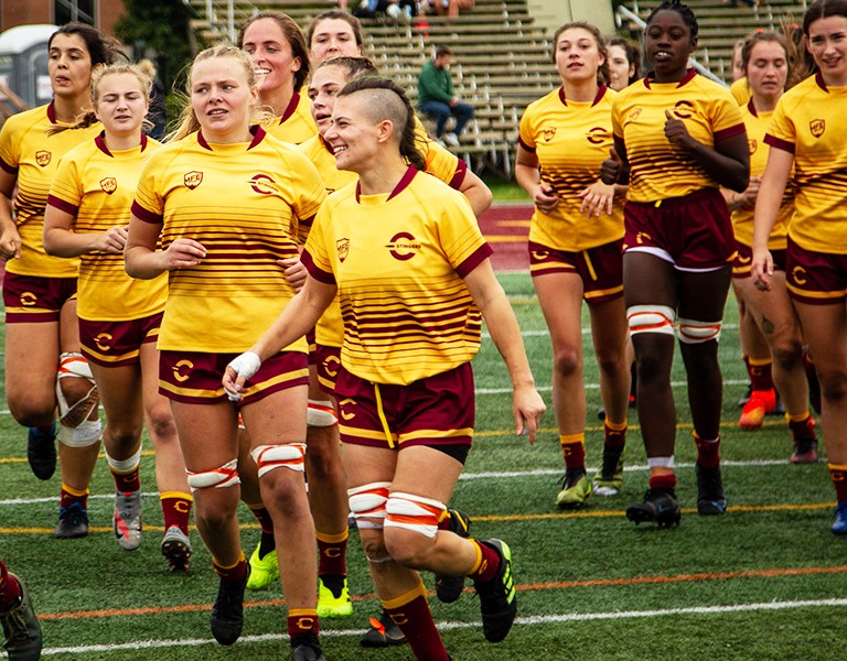 A group of women rugby players in a yellow and burgundy uniforms.