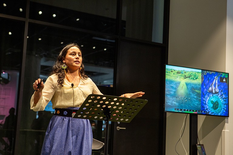 The inaugural Miywâcimo! storytelling competition was held at 4TH SPACE in March of this year. Young woman with long, dark hair, standing behind a lectern and looking out to an audience.