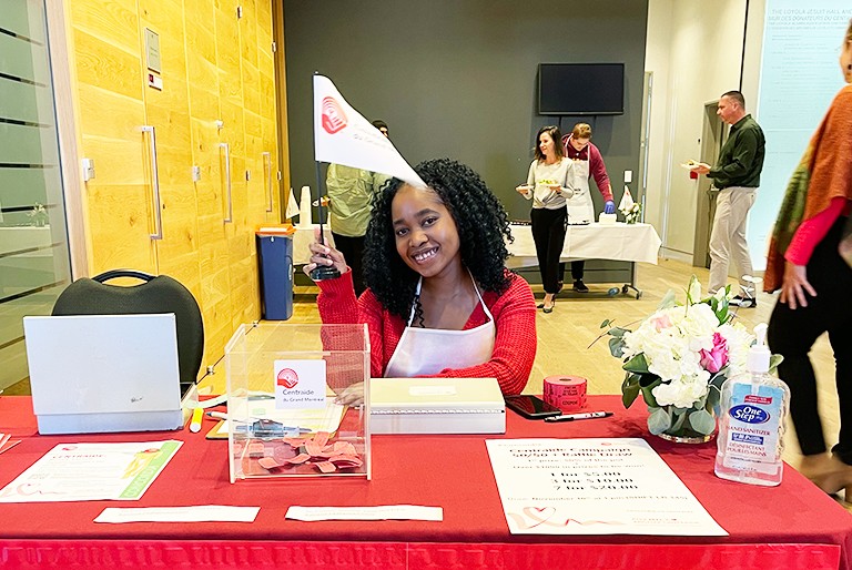 Young, smiling women with long, dark, curly hair, wearing a red long-sleeved top and a white apron and holding a white flag with the Centraide logo.