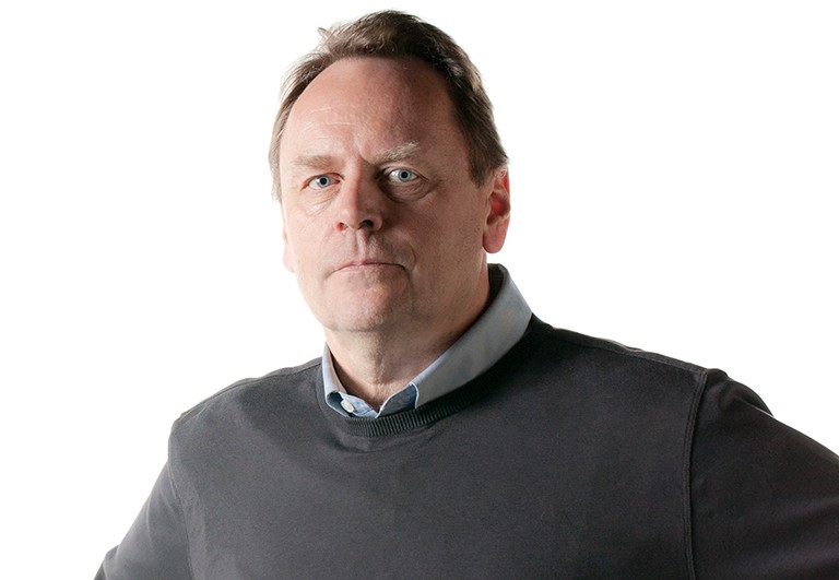 Middle aged man with blue eyes and wearing a blue collared shirt and grey sweater, looking seriously at the camera