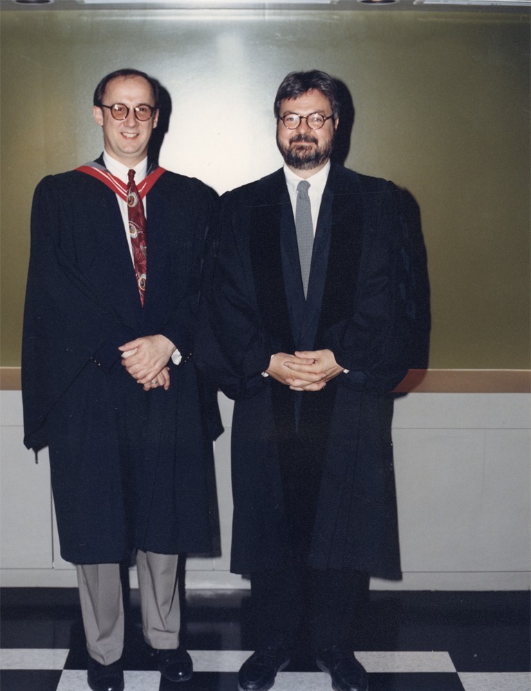 Two men in university regalia pose for a photo, hands clasped in front of them while smiling.