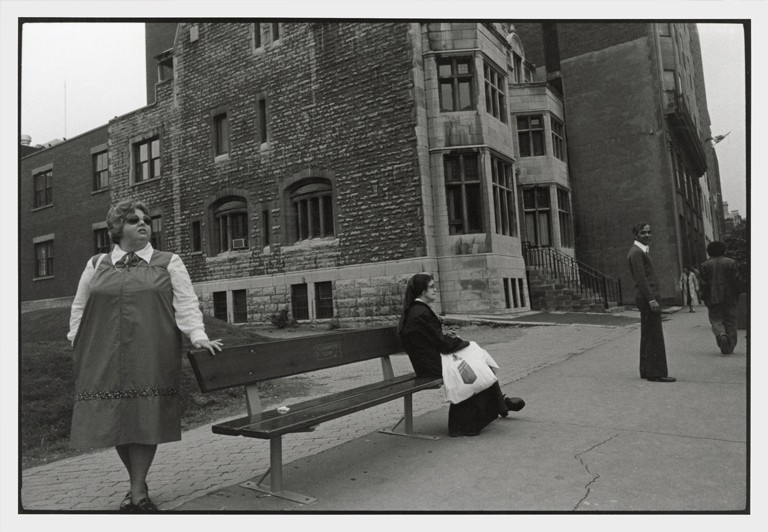 A woman leans against a bench that a nun is sitting on. An old building sits in the background.