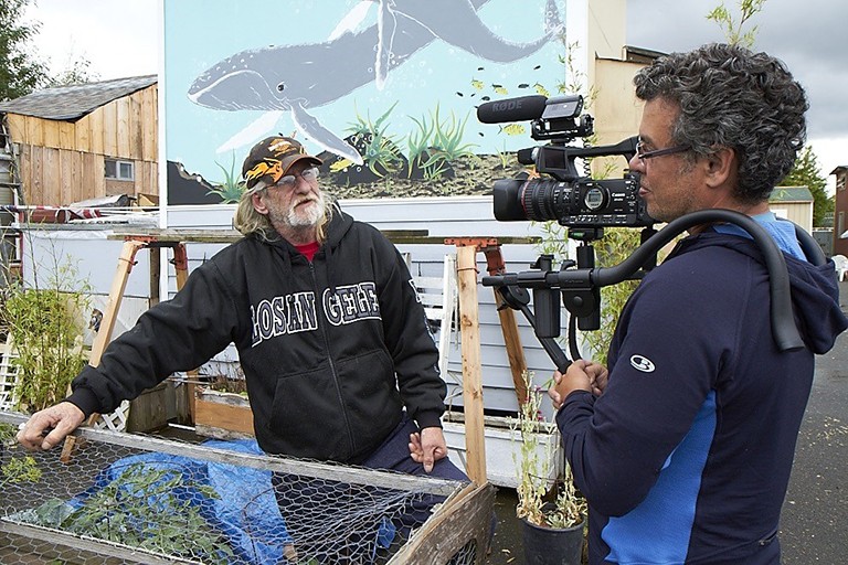 An outdoor urban garden where a man (right) with short, greying hair holds a camera on his shoulder and talks to a man with long, greying blonde hair, glasses and a beard.