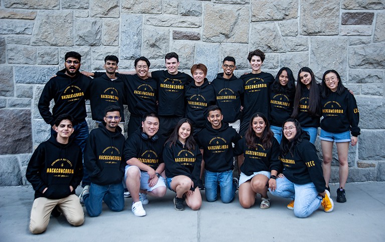 A group of smiling young people in black hooded sweatshirts that have the words "HackConcordia" in yellow writing on the front.