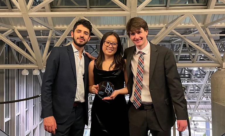 Two young men and one woman, wearing formal wear and standing together, with the woman holding an award shaped out of a round, glass disk.