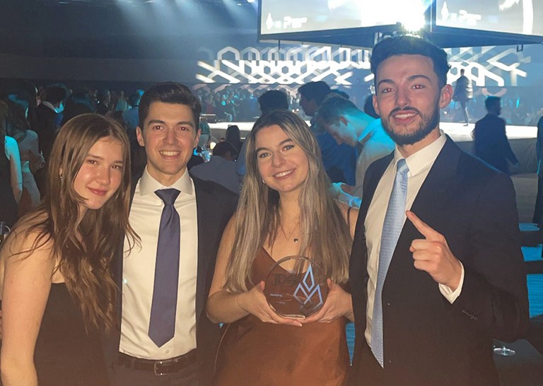 A group of young people (two women and two men) wearing formal wear and standing together in a crowded event centre.