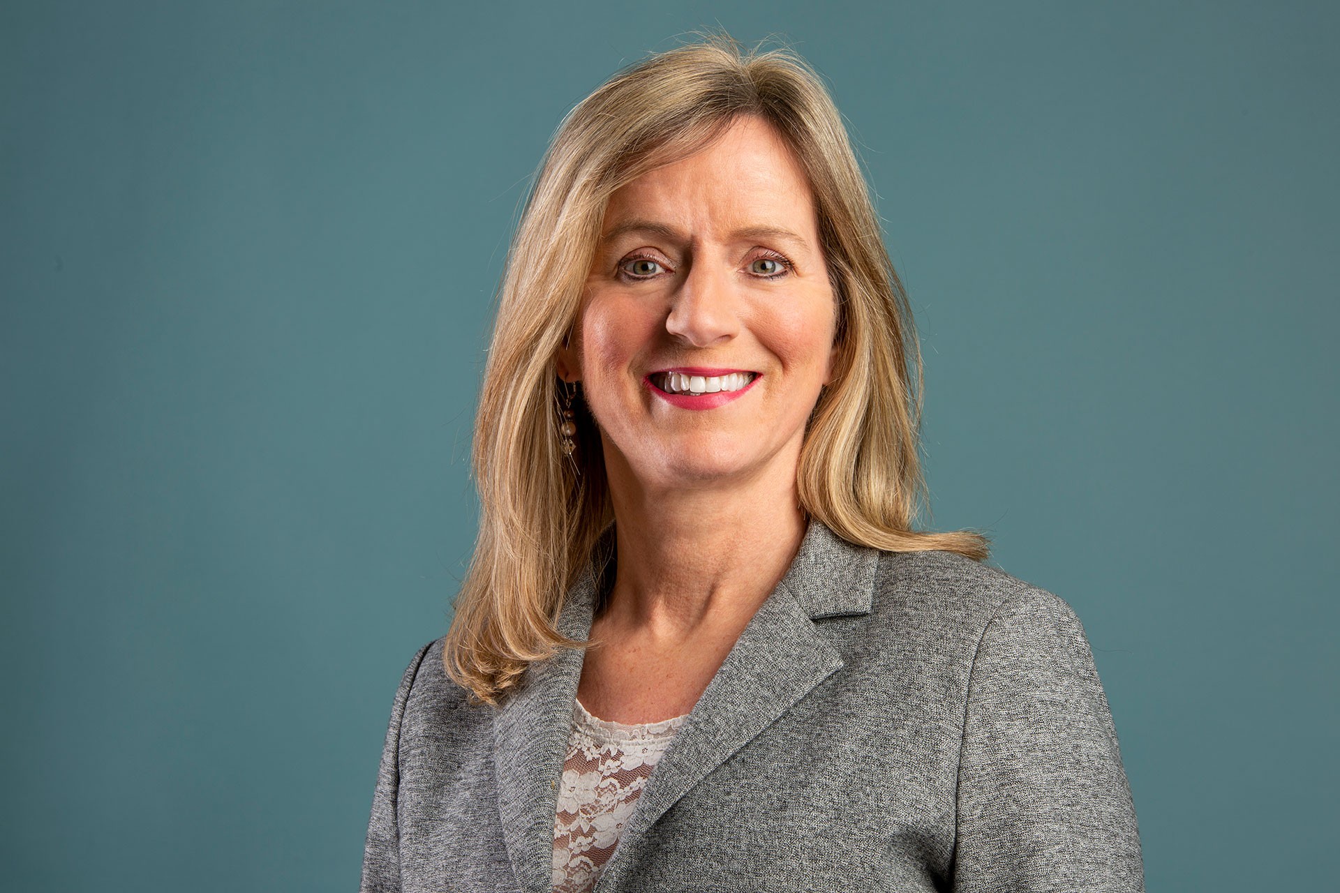 Smiling woman with bright pink lipstick, long blonde hair and wearing a pale knit turtleneck and a blue blazer.