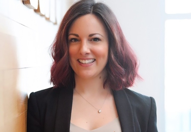 Smiling woman with long, dark hair, wearing a gold necklace, beige top and black blazer.