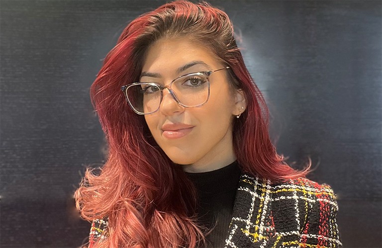 Smiling woman with glasses and long red hair, wearing a black top and a patterned blazer.