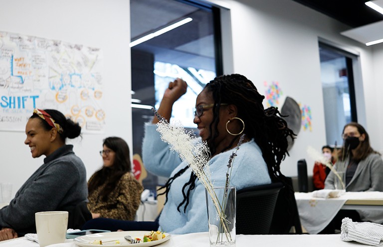 Ushana Houston: “Many of these families are facing structural barriers. We are there to accompany them.” | Photo: Marta Mostardini Smiling women sitting in a room at several long tables and facing the front with one woman in the foreground raising her hand.